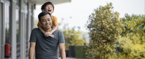 Father carrying his laughing son on his back, walking outdoors on a sunny day.